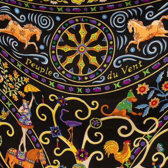 A close-up of a black textile scarf featuring a highly detailed, colorful folk-art print with horses, parrots, a rooster, floral motifs, and swirling gold lines. The text 'Peuple du Vent' is printed across the center wheel.