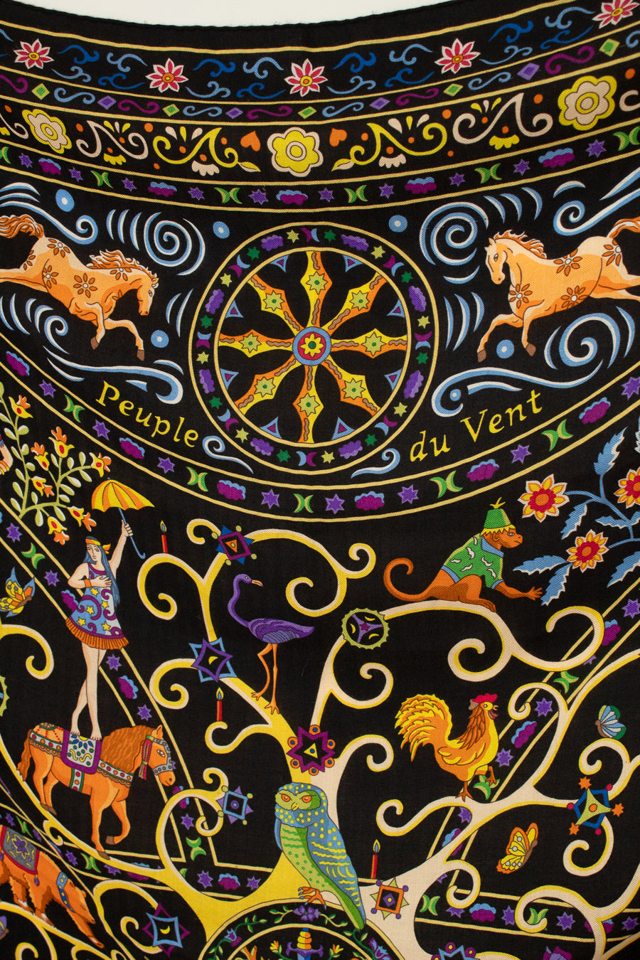 A close-up of a black textile scarf featuring a highly detailed, colorful folk-art print with horses, parrots, a rooster, floral motifs, and swirling gold lines. The text 'Peuple du Vent' is printed across the center wheel.