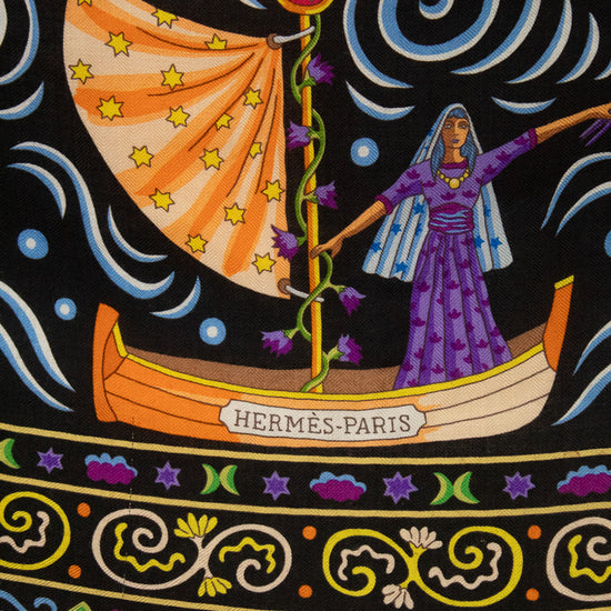 A close-up of a black textile scarf featuring a highly detailed, colorful folk-art print with horses, parrots, a rooster, floral motifs, and swirling gold lines. The text 'Hermes-Paris' printed across the center.