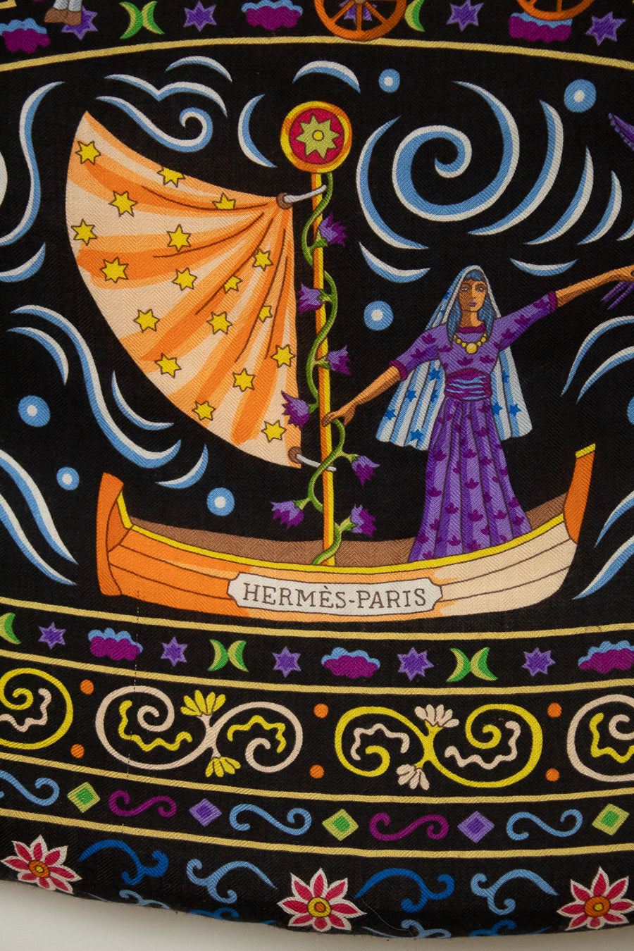 A close-up of a black textile scarf featuring a highly detailed, colorful folk-art print with horses, parrots, a rooster, floral motifs, and swirling gold lines. The text 'Hermes-Paris' printed across the center.