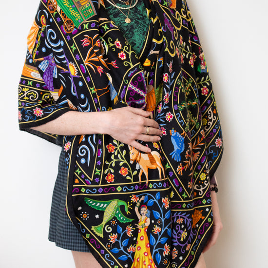 A model wearing a black silk scarf with a vibrant, multicolored bohemian print featuring peacocks, flowers, and traditional patterns, styled over a black top and a dark plaid skirt against a plain white background.