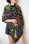 A model wearing a black silk scarf with a vibrant, multicolored bohemian print featuring peacocks, flowers, and traditional patterns, styled over a black top and a dark plaid skirt against a plain white background.
