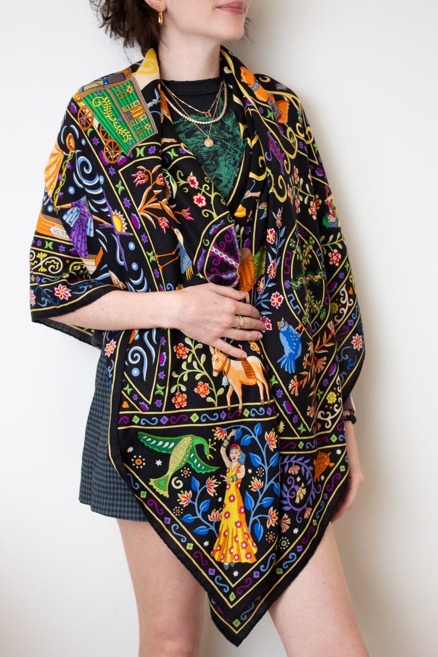A model wearing a black silk scarf with a vibrant, multicolored bohemian print featuring peacocks, flowers, and traditional patterns, styled over a black top and a dark plaid skirt against a plain white background.