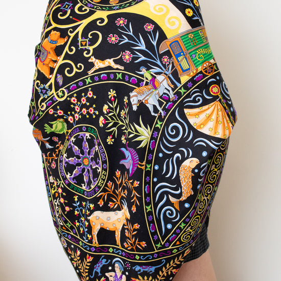 A model wearing a black silk scarf with a vibrant, multicolored bohemian print featuring peacocks, flowers, and traditional patterns, against a plain white background.