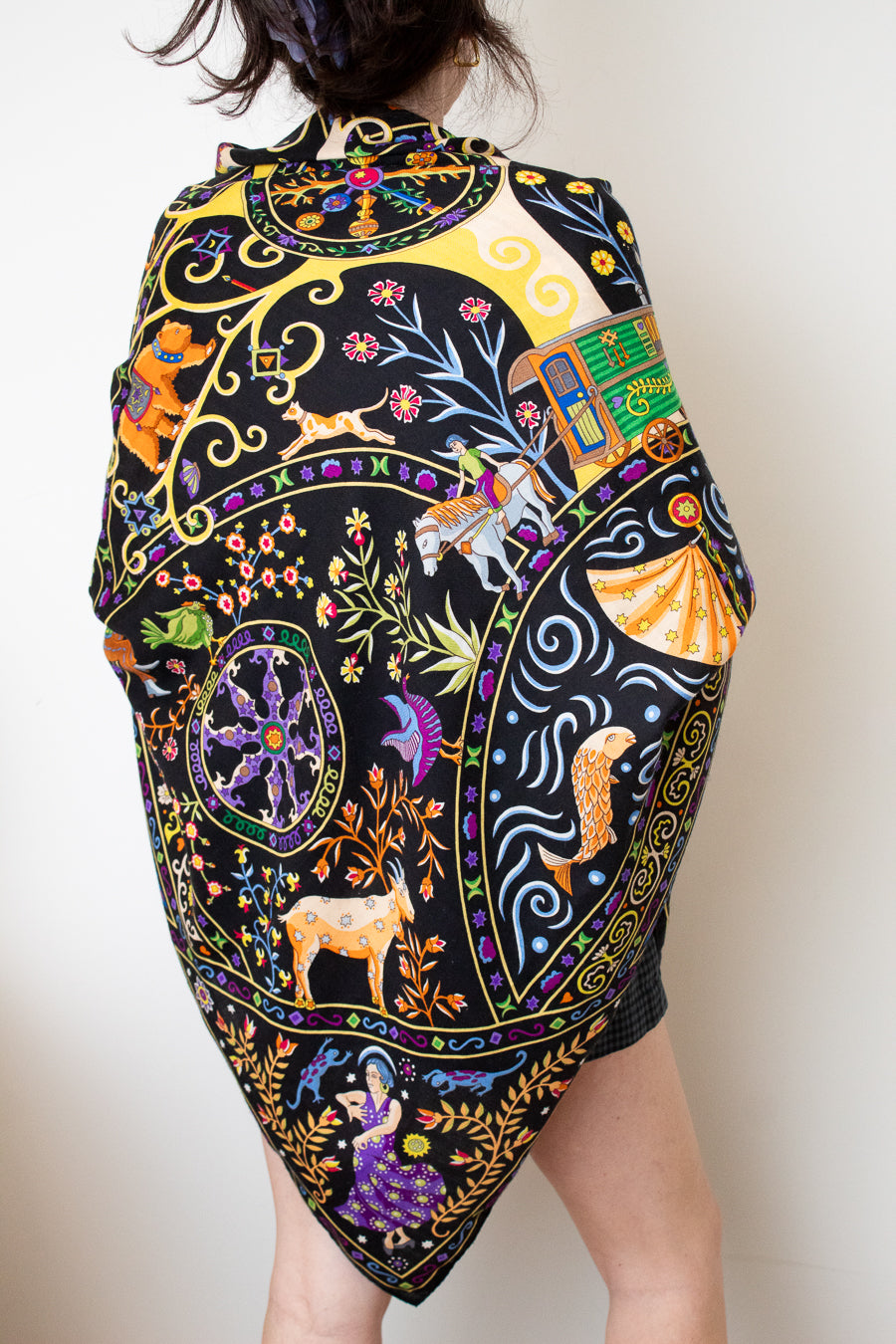 A model wearing a black silk scarf with a vibrant, multicolored bohemian print featuring peacocks, flowers, and traditional patterns, against a plain white background.
