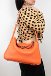 An orange pebbled leather shoulder bag with silver hardware, worn by a model wearing a beige and black polka dot blouse against a plain white background.