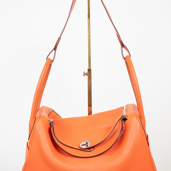 An orange pebbled leather shoulder bag with a curved top, silver-tone hardware, and a matching orange leather strap, displayed on a white background with a gold-colored stand.
