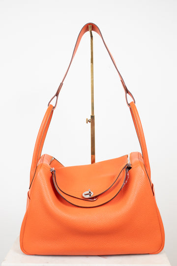 An orange pebbled leather shoulder bag with a curved top, silver-tone hardware, and a matching orange leather strap, displayed on a white background with a gold-colored stand.