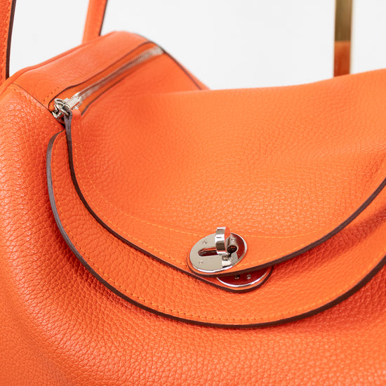 Close-up of an orange pebbled leather handbag with a silver-tone turn-lock closure and dark contrast stitching on a light gray background.