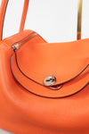 Close-up of an orange pebbled leather handbag with a silver-tone turn-lock closure and dark contrast stitching on a light gray background.