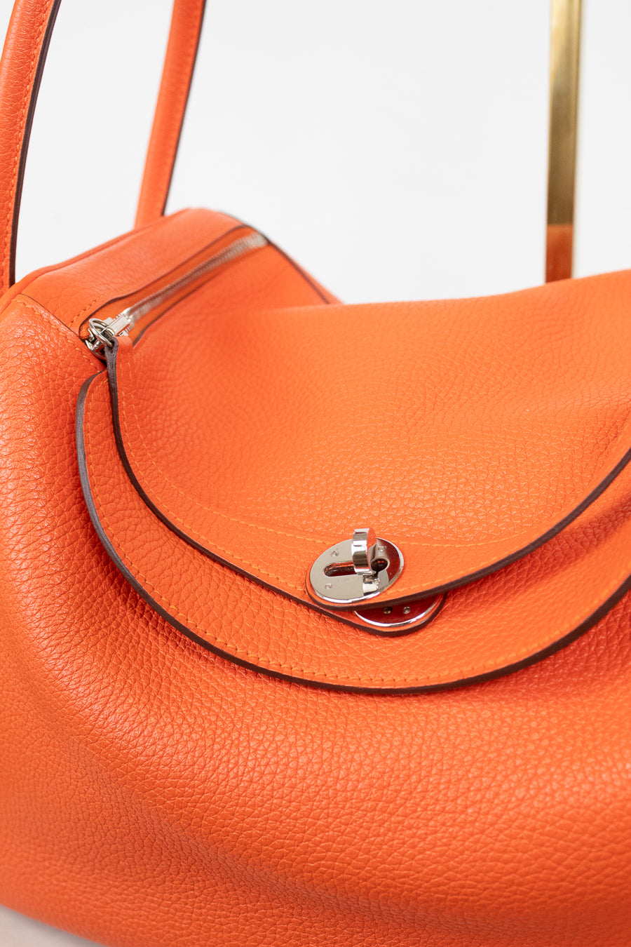 Close-up of an orange pebbled leather handbag with a silver-tone turn-lock closure and dark contrast stitching on a light gray background.