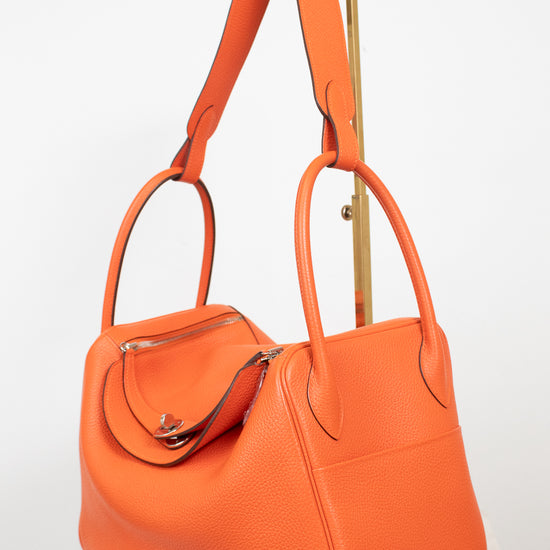 An orange pebbled leather handbag with dual top handles and a shoulder strap, displayed on a white marble pedestal against a white background.