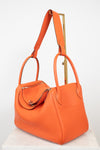 An orange pebbled leather handbag with dual top handles and a shoulder strap, displayed on a white marble pedestal against a white background.