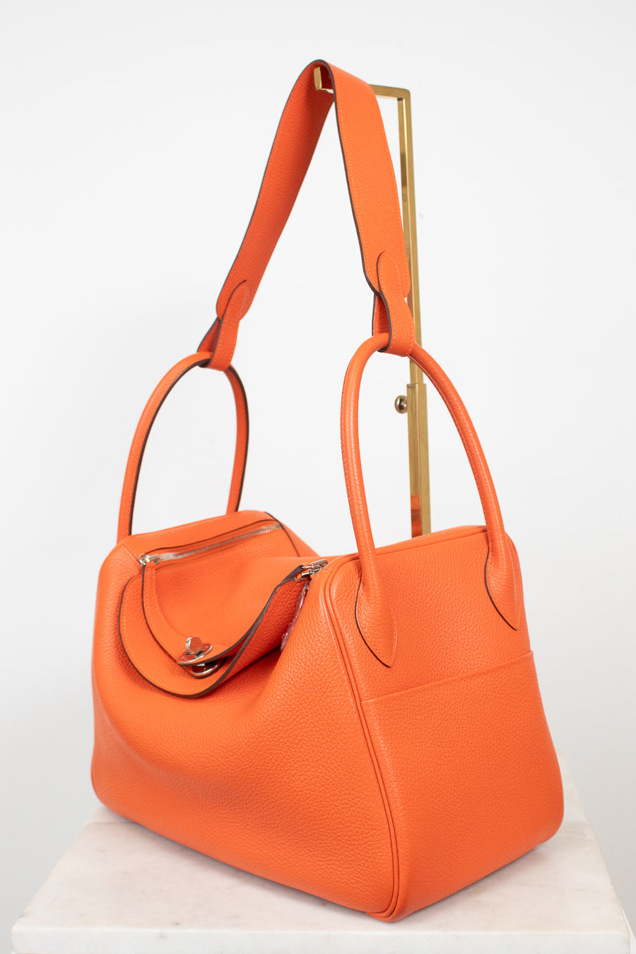An orange pebbled leather handbag with dual top handles and a shoulder strap, displayed on a white marble pedestal against a white background.
