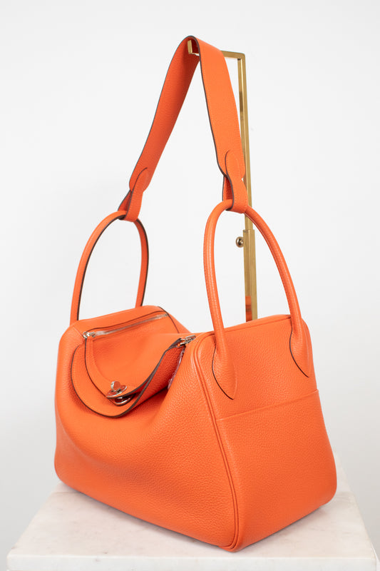 An orange pebbled leather handbag with dual top handles and a shoulder strap, displayed on a white marble pedestal against a white background.