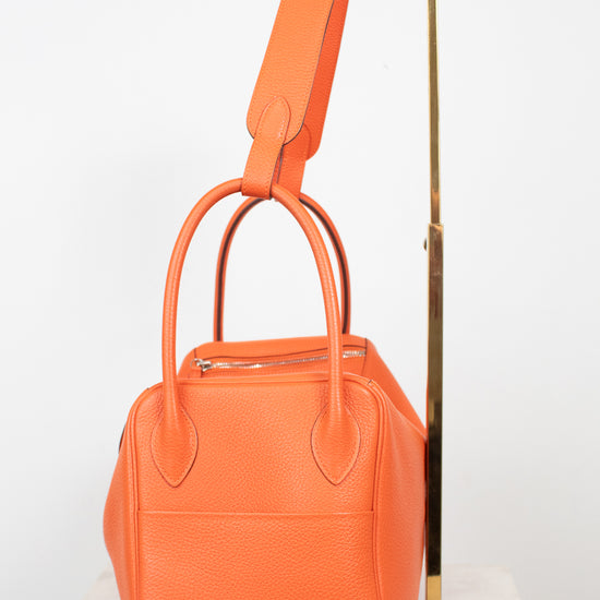 An orange textured leather handbag with double top handles and a matching shoulder strap, displayed on a white marble surface against a white background with a gold metal stand.