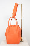 An orange textured leather handbag with double top handles and a matching shoulder strap, displayed on a white marble surface against a white background with a gold metal stand.