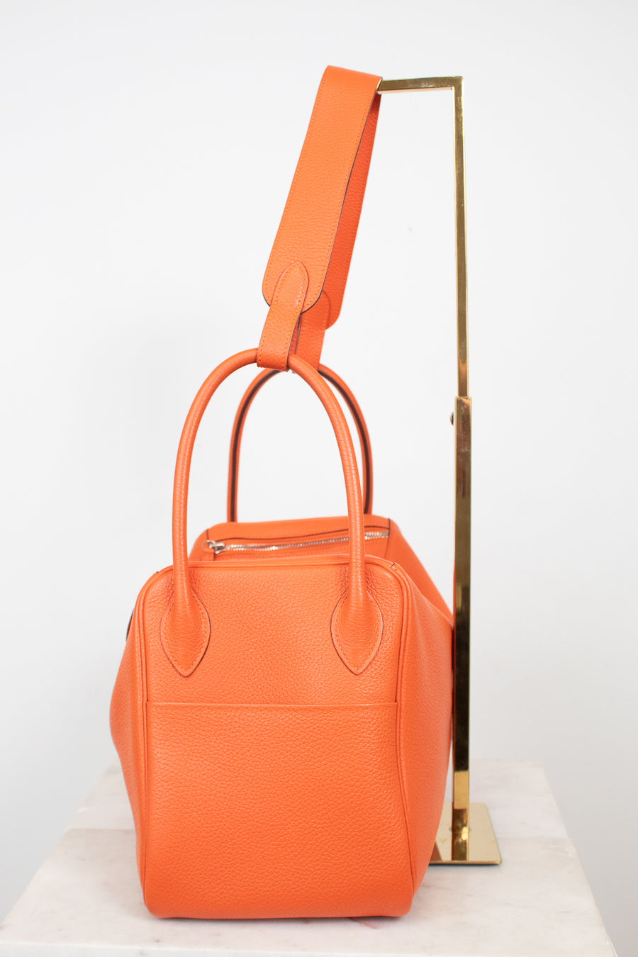 An orange textured leather handbag with double top handles and a matching shoulder strap, displayed on a white marble surface against a white background with a gold metal stand.