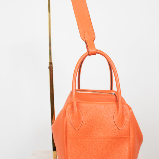 An orange pebbled leather handbag with dual top handles and a matching shoulder strap, displayed on a white marble surface next to a gold metal stand against a white background.