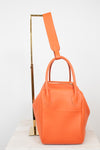 An orange pebbled leather handbag with dual top handles and a matching shoulder strap, displayed on a white marble surface next to a gold metal stand against a white background.