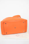 Bright orange pebbled leather handbag shown from the bottom, featuring silver-tone metal studs and a matching orange strap, set against a plain white background.