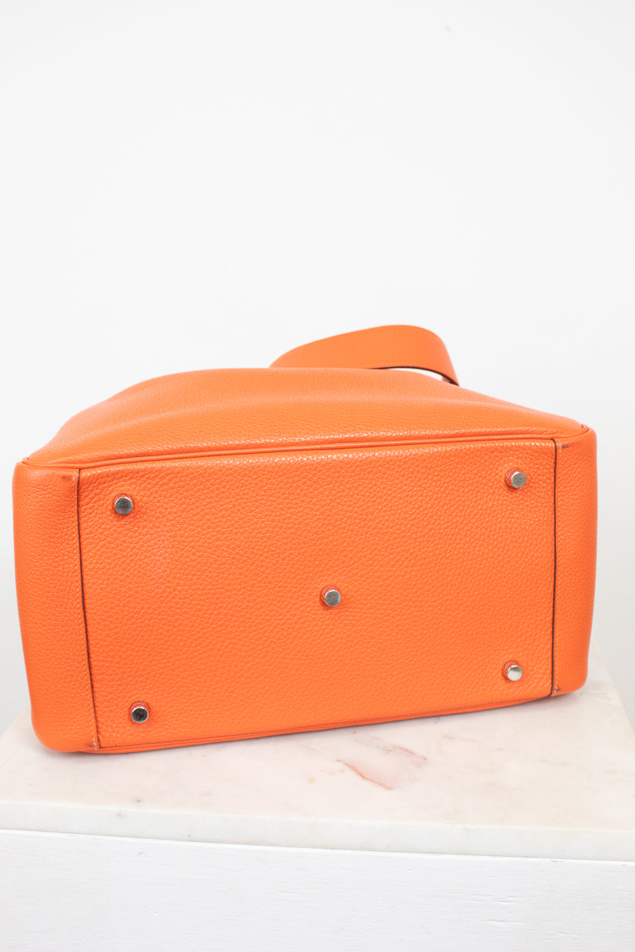 Bright orange pebbled leather handbag shown from the bottom, featuring silver-tone metal studs and a matching orange strap, set against a plain white background.