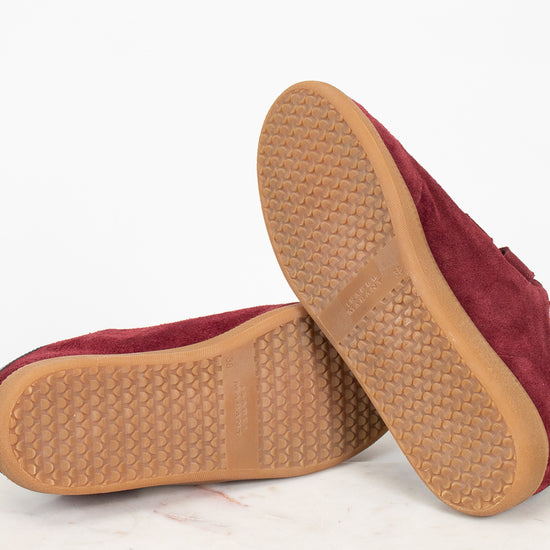 A pair of maroon suede shoes shown from the bottom, highlighting the tan textured rubber soles, set against a clean white background.