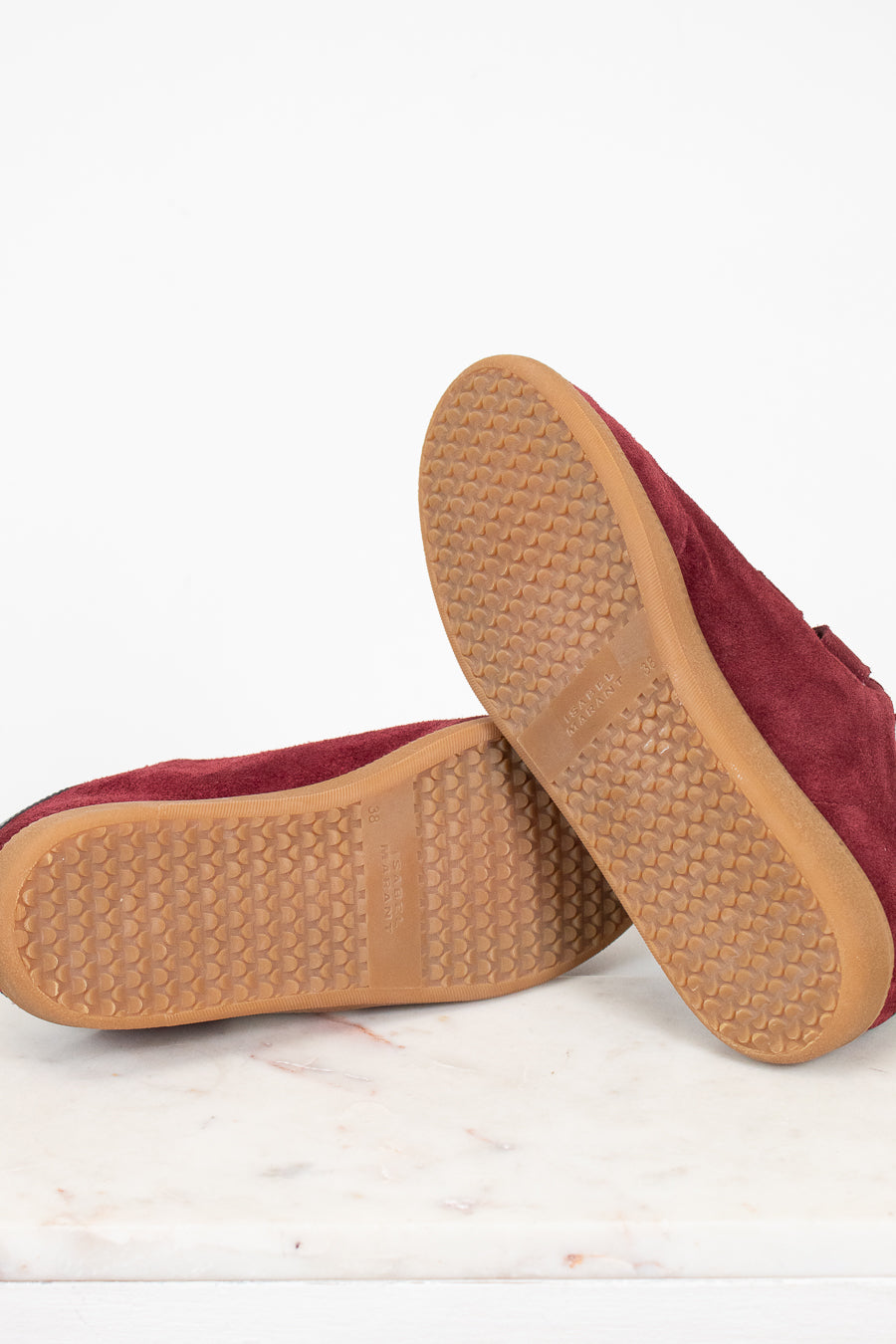 A pair of maroon suede shoes shown from the bottom, highlighting the tan textured rubber soles, set against a clean white background.