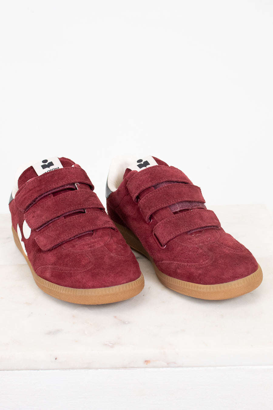 A pair of maroon suede shoes with triple Velcro straps and gum soles, displayed on a light marble surface against a white background.