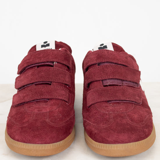 A pair of maroon suede sneakers with triple Velcro straps and gum soles, photographed from the front on a light marble surface against a white background.