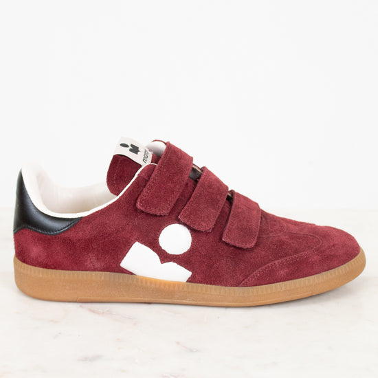 A low-top sneaker in maroon suede with triple Velcro straps, a black heel tab, a white side logo, and a tan rubber sole, photographed on a white marble surface against a white background.
