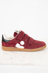 A low-top sneaker in maroon suede with triple Velcro straps, a black heel tab, a white side logo, and a tan rubber sole, photographed on a white marble surface against a white background.