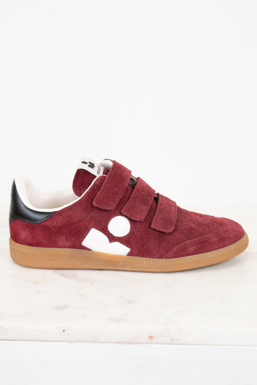 A low-top sneaker in maroon suede with triple Velcro straps, a black heel tab, a white side logo, and a tan rubber sole, photographed on a white marble surface against a white background.