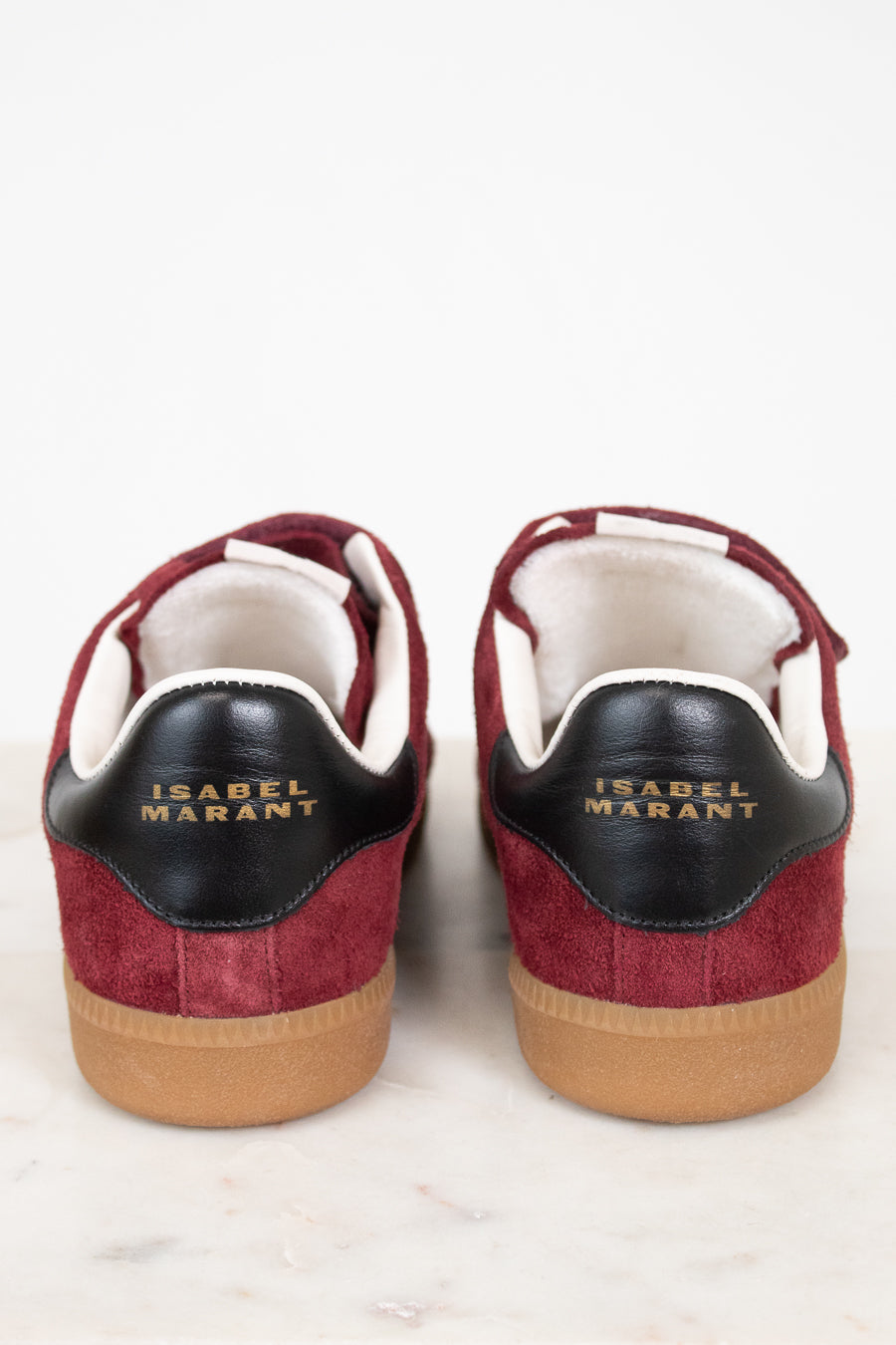 A pair of Isabel Marant sneakers shown from the back, featuring burgundy suede and black leather details with gold lettering, white lining, and tan rubber soles, set against a clean white background.