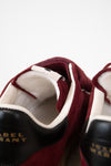 Close-up of a pair of suede sneakers in burgundy and black with white lining and gold 'ISABEL MARANT' branding on the tongue.