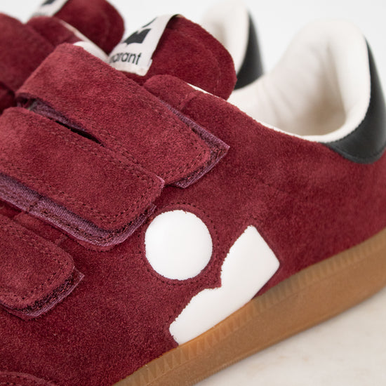A close-up of a pair of suede sneakers in maroon with white graphic side logos, triple maroon Velcro straps, a white interior lining, and a brown gum sole.