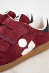 A close-up of a pair of suede sneakers in maroon with white graphic side logos, triple maroon Velcro straps, a white interior lining, and a brown gum sole.
