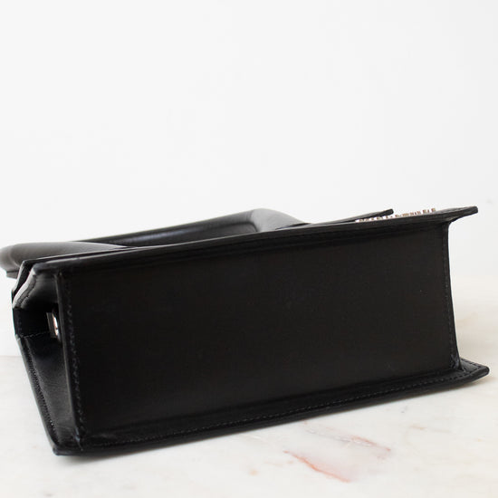 Bottom view of a black leather bag with a structured rectangular shape, resting on a white marble surface against a white background.
