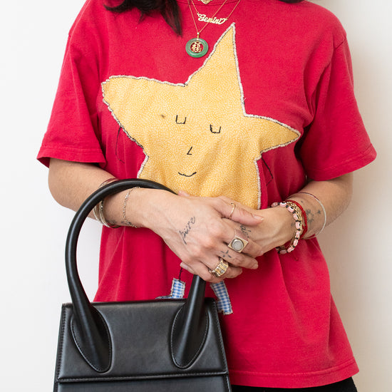 A woman wearing a red graphic t-shirt with a large yellow star design, holding a black structured handbag. She is also wearing gold jewelry and black shorts, set against a plain off-white background.