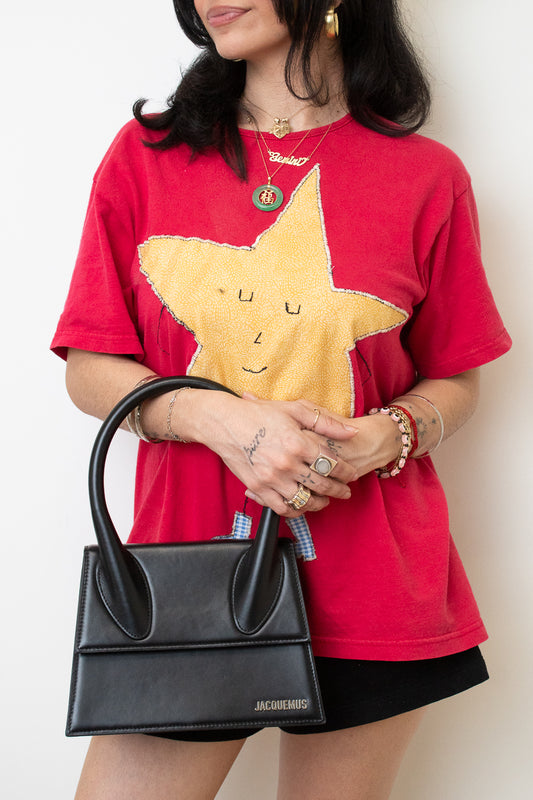 A woman wearing a red graphic t-shirt with a large yellow star design, holding a black structured handbag. She is also wearing gold jewelry and black shorts, set against a plain off-white background.