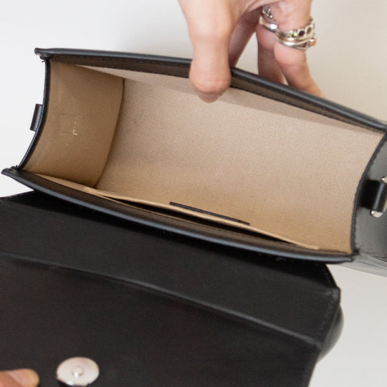 A close-up of a black leather handbag being opened to show a beige interior lining, with hands holding the flap and a silver snap button visible.