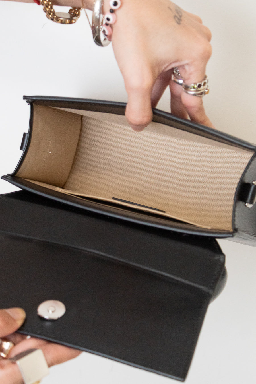 A close-up of a black leather handbag being opened to show a beige interior lining, with hands holding the flap and a silver snap button visible.