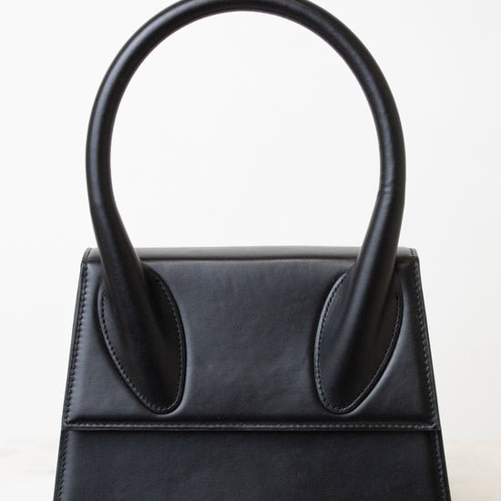 A black leather handbag with a distinctive top handle, featuring a structured rectangular body and the brand name 'JACQUEMUS' in silver lettering on the bottom right, set against a clean white background.