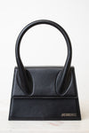 A black leather handbag with a distinctive top handle, featuring a structured rectangular body and the brand name 'JACQUEMUS' in silver lettering on the bottom right, set against a clean white background.