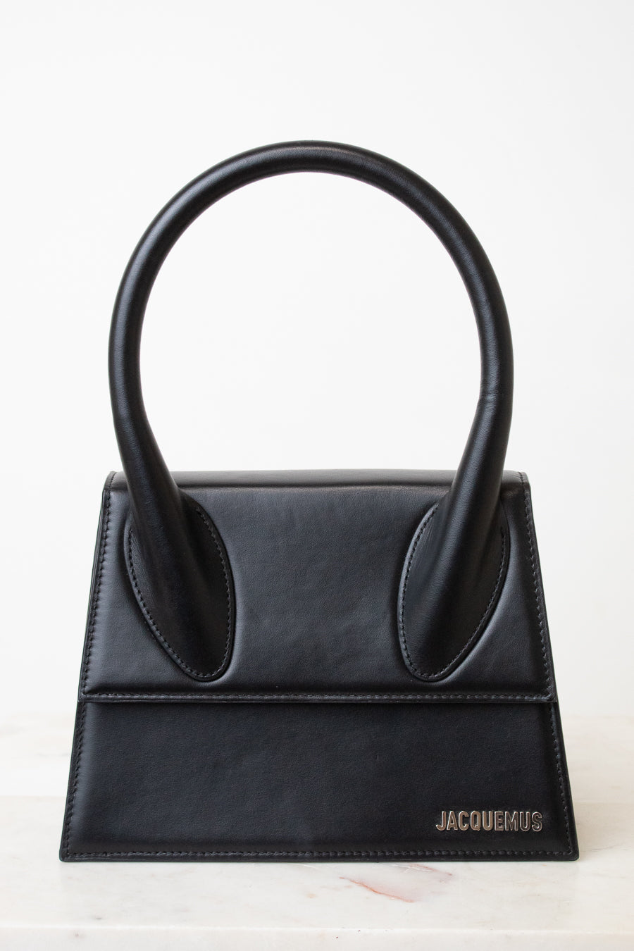 A black leather handbag with a distinctive top handle, featuring a structured rectangular body and the brand name 'JACQUEMUS' in silver lettering on the bottom right, set against a clean white background.