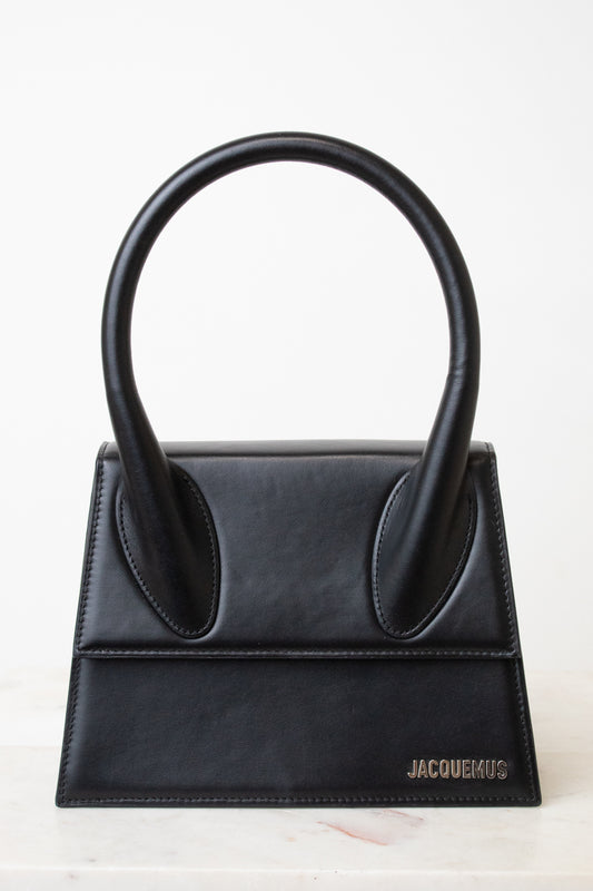 A black leather handbag with a distinctive top handle, featuring a structured rectangular body and the brand name 'JACQUEMUS' in silver lettering on the bottom right, set against a clean white background.