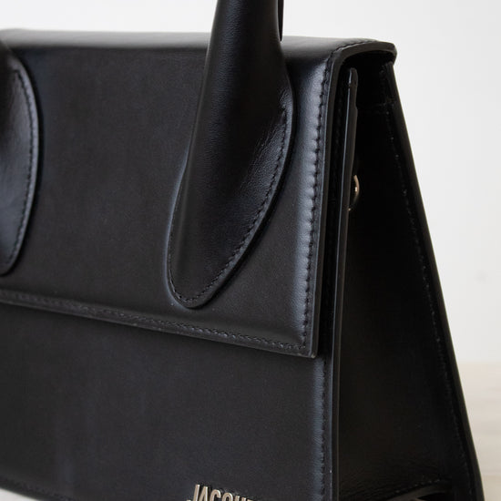 A close-up of a black leather handbag by Jacquemus, showing the side flap, top handle, and the brand name in silver lettering on the front, set against a light neutral background.