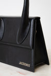 A close-up of a black leather handbag by Jacquemus, showing the side flap, top handle, and the brand name in silver lettering on the front, set against a light neutral background.