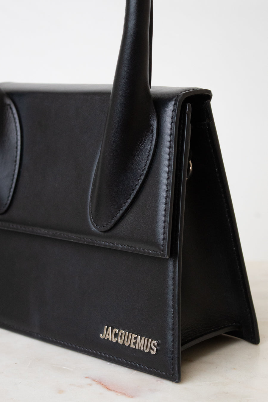 A close-up of a black leather handbag by Jacquemus, showing the side flap, top handle, and the brand name in silver lettering on the front, set against a light neutral background.