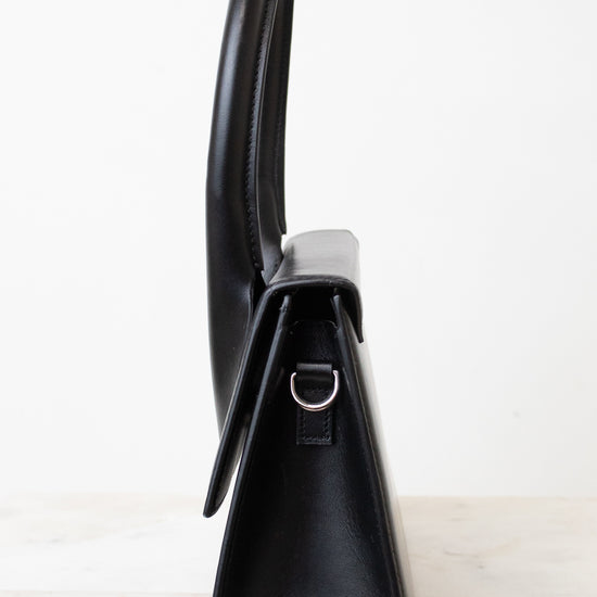 A black leather shoulder bag shown from a side profile view, standing upright on a light marble surface against a white background.